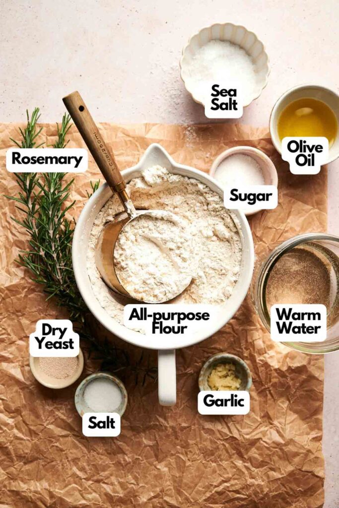 Ingredients for focaccia bread are arranged on a brown paper surface, including rosemary, all-purpose flour, sea salt, olive oil, sugar, dry yeast, salt, warm water, and garlic.