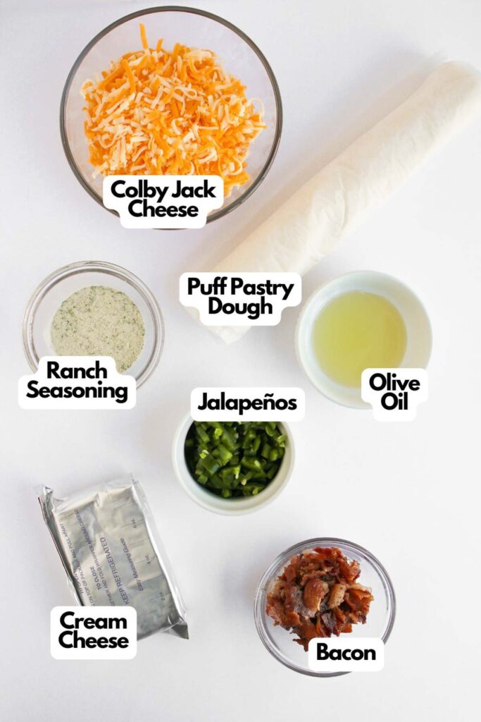 Top view of ingredients for jalapeno popper pinwheels, including Colby Jack cheese, puff pastry dough, olive oil, Ranch seasoning, jalapeños, bacon, and cream cheese, all artfully arranged on a white surface.