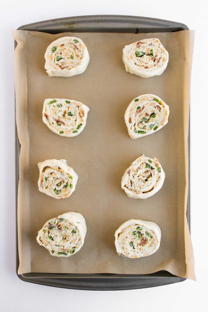 Eight unbaked jalapeno popper pinwheels with a cream and green filling rest on a parchment-lined baking tray.