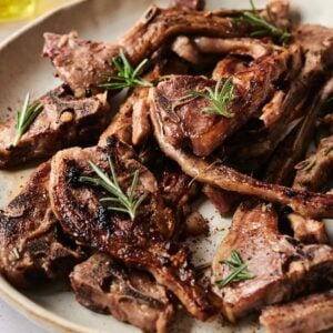 Succulent grilled lamb chops rest on a beige plate, beautifully garnished with sprigs of rosemary.