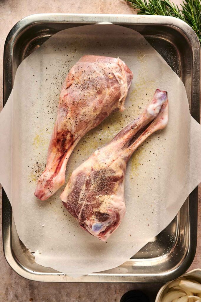Two raw lamb shanks, reminiscent of a succulent leg of lamb, seasoned with salt and pepper on parchment paper in a metal baking tray, ready for cooking.