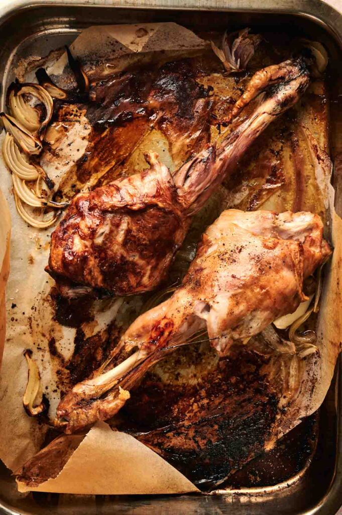 Two roasted lamb shanks, reminiscent of a succulent leg of lamb, rest on a baking tray lined with parchment paper, surrounded by tender cooked onions.