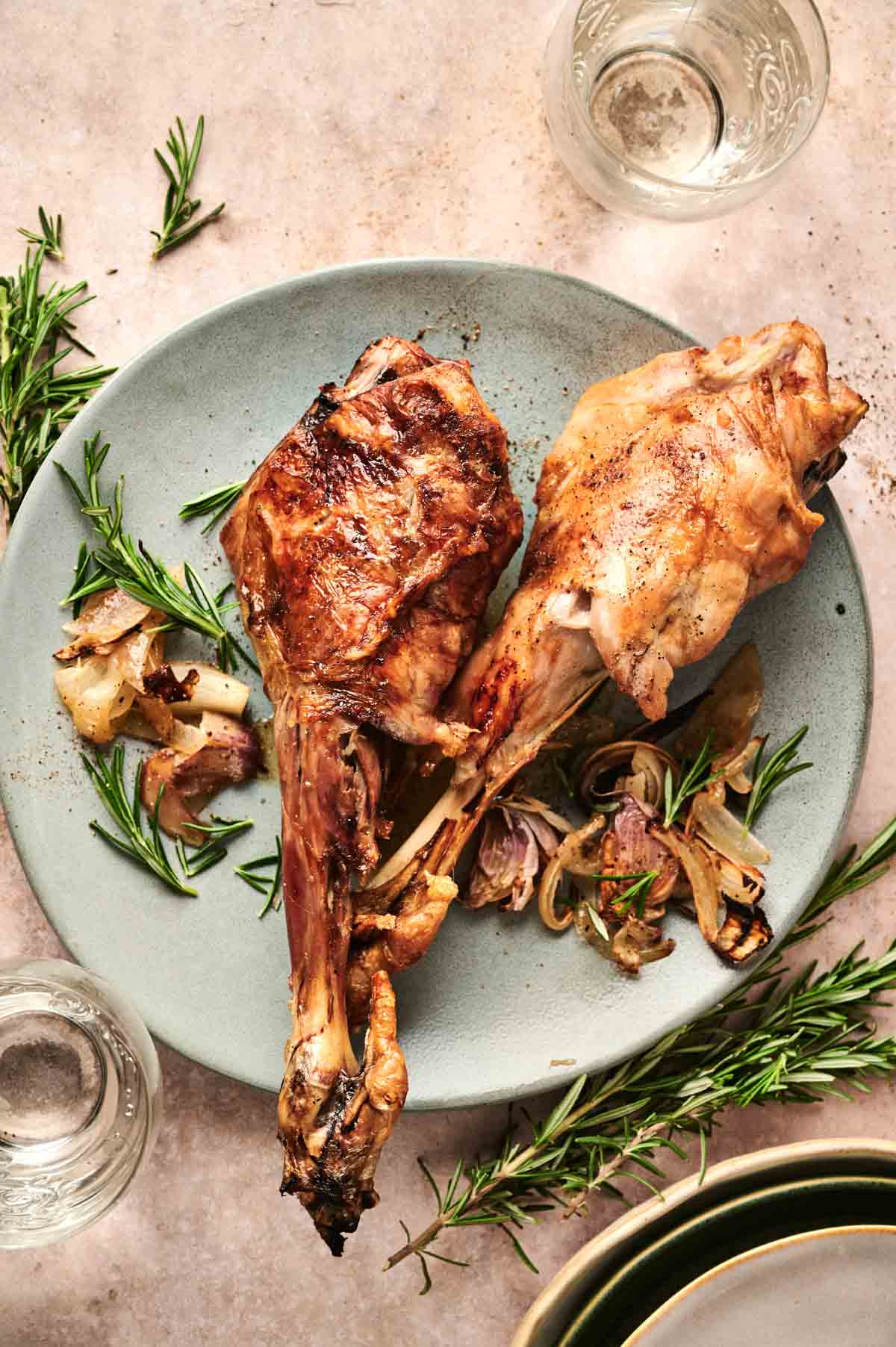 Two roasted lamb shanks resembling a tender leg of lamb sit on a plate with cooked onions and rosemary sprigs, surrounded by empty glasses and more rosemary, set against a neutral background.