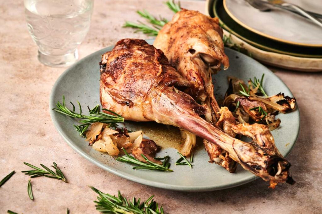 Two succulent roasted lamb shanks, reminiscent of a classic leg of lamb, are presented with aromatic herbs on a plate, elegantly garnished with rosemary sprigs. A glass and neatly stacked dishes complete the inviting scene.