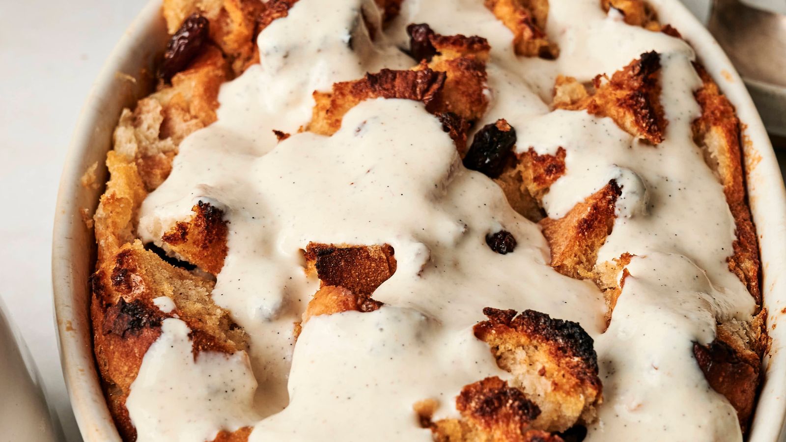 A close-up of bread pudding topped with creamy vanilla sauce in a white baking dish. The pudding features toasted bread pieces and visible raisins.