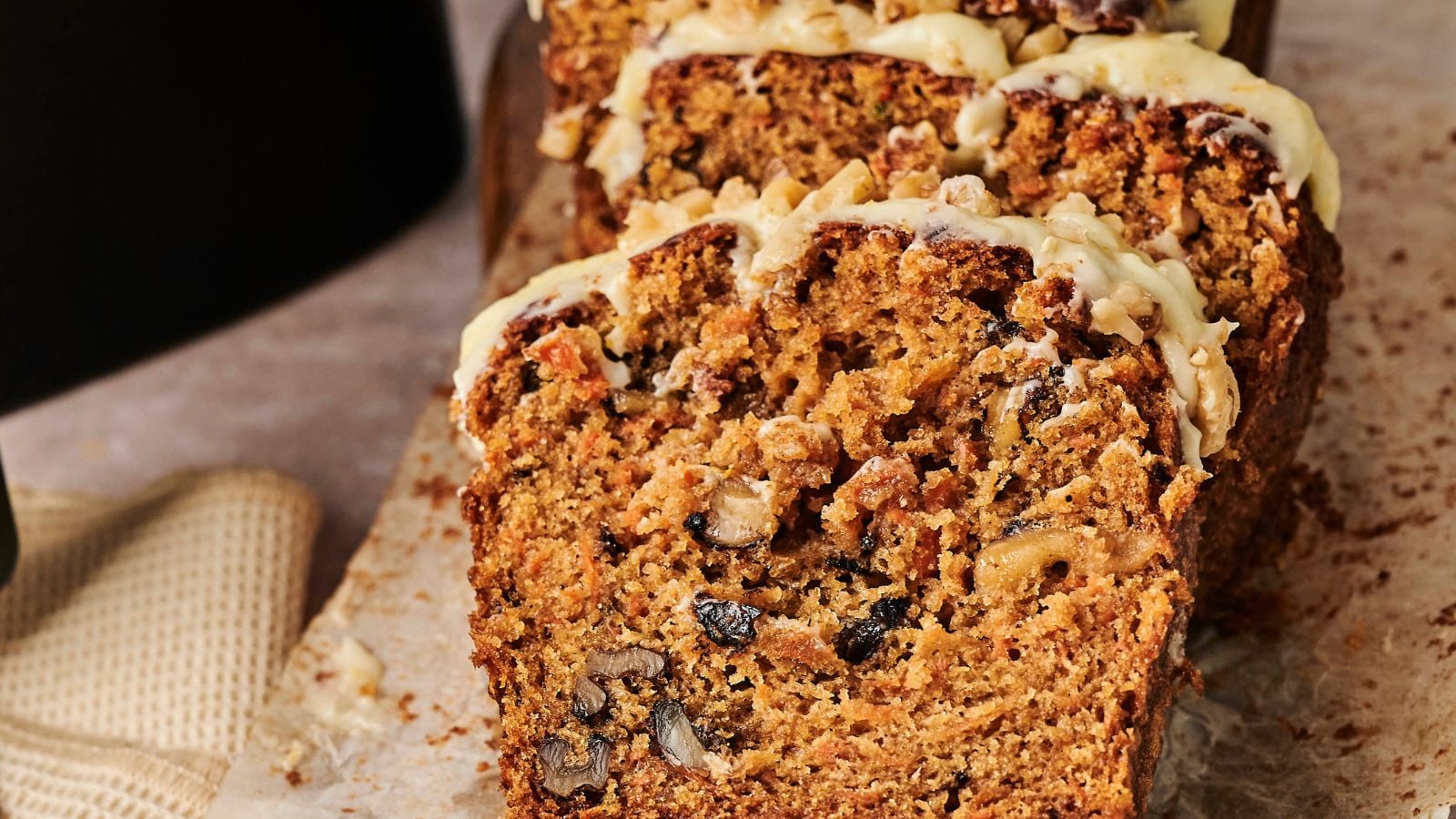 Sliced carrot cake with nuts is topped with cream cheese frosting, placed on a piece of parchment paper next to a napkin.