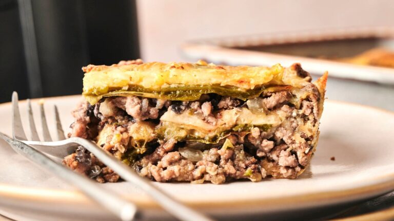 A slice of moussaka on a plate with a fork, featuring layered ground meat, eggplant, and a creamy topping.