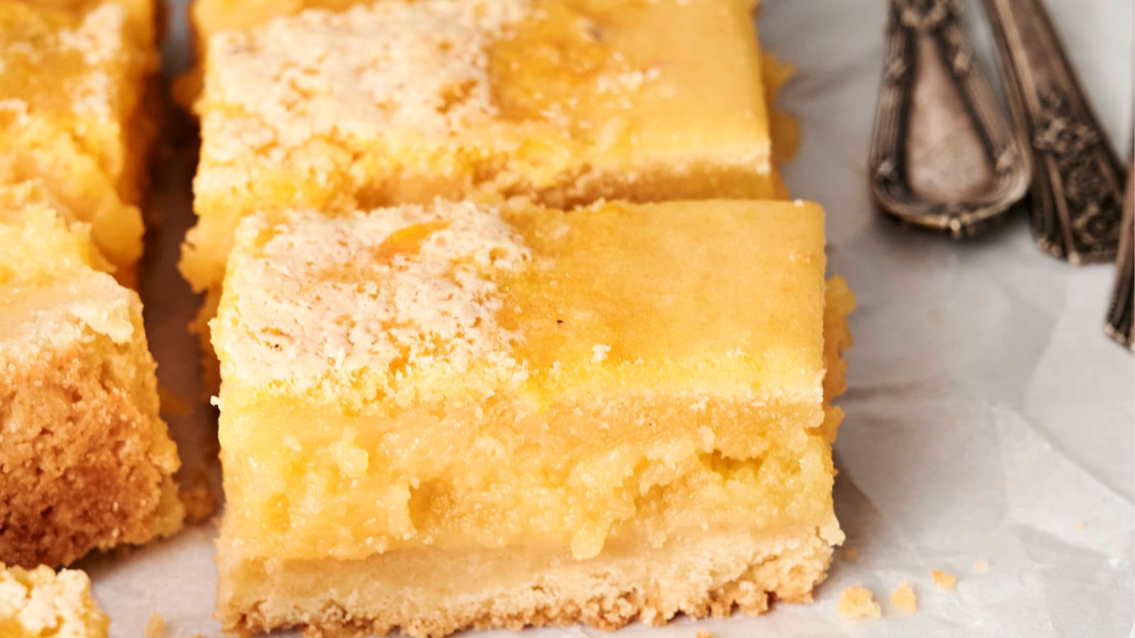 Close-up of lemon bars with a crumbly base and a smooth, yellow filling, cut into squares, placed on parchment paper. Silver utensils are visible to the side.