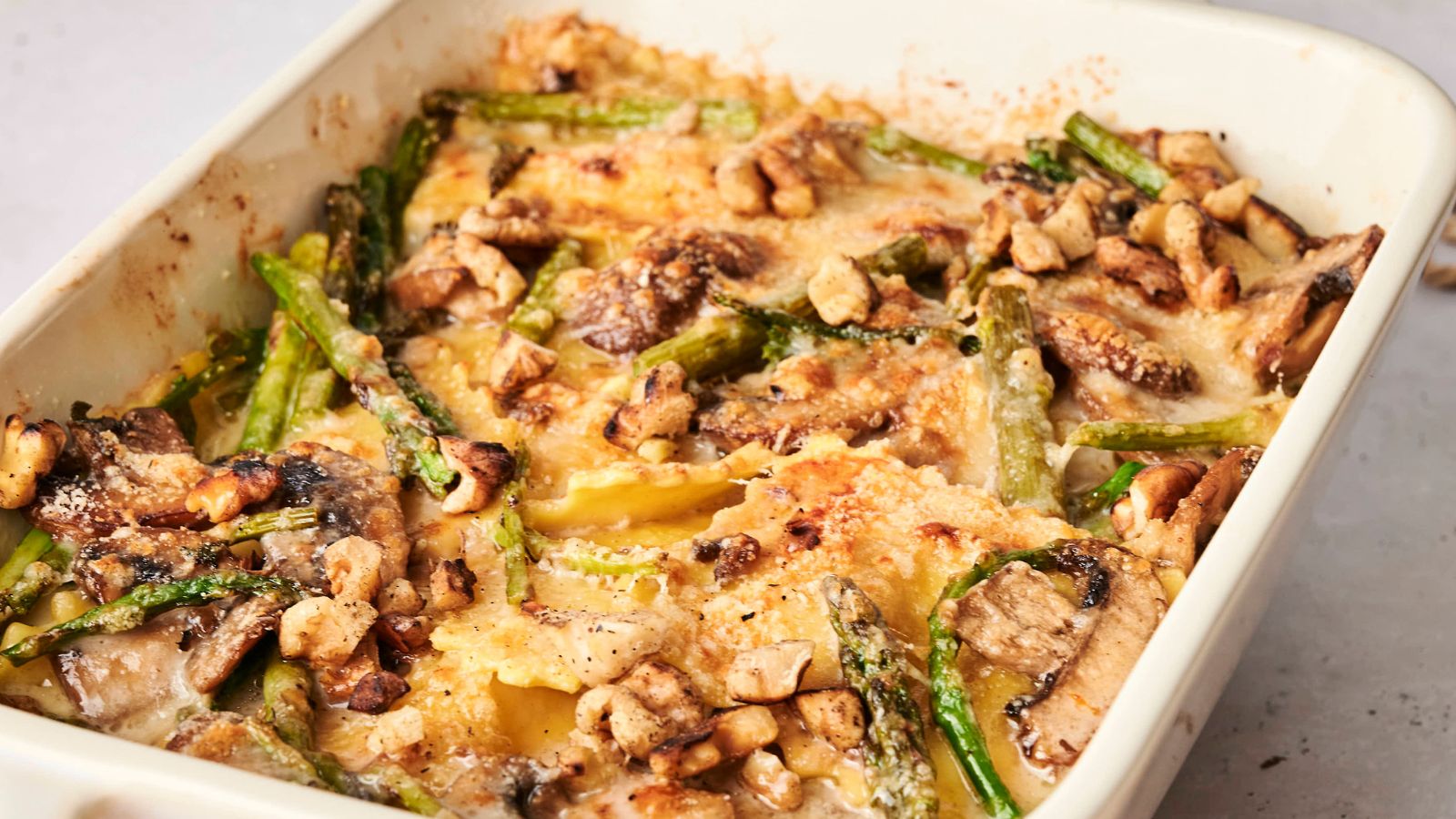 Baked dish with layered pasta, mushrooms, asparagus, and nuts in a creamy sauce, topped with a golden-brown crust, in a rectangular white baking dish.