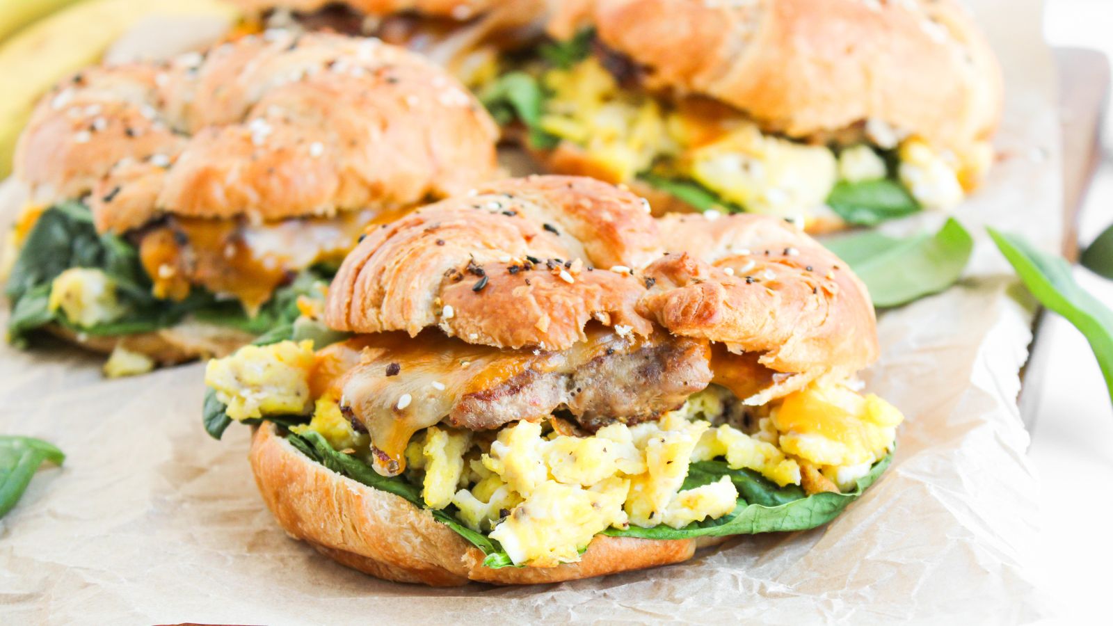 Croissant sandwich with scrambled eggs, sausage patty, melted cheese, and spinach on parchment paper.