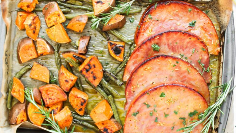 Slices of cooked ham with parsley, roasted sweet potatoes, green beans, and rosemary sprigs on a baking tray.