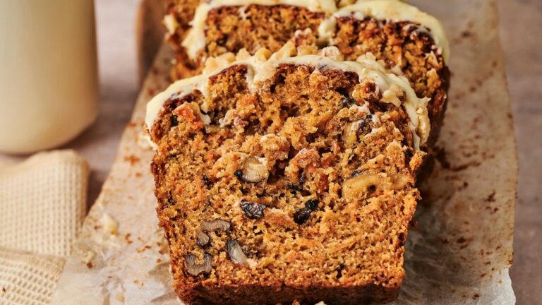 Close-up of sliced carrot cake on parchment paper, showing a moist texture with visible raisins, walnuts, and a light drizzle of icing on top.