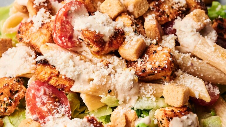 Close-up of a Caesar salad with lettuce, grilled chicken pieces, penne pasta, cherry tomatoes, croutons, Caesar dressing, and grated cheese.