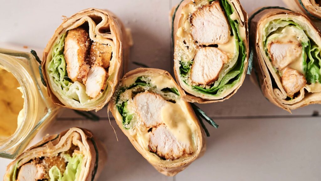 Chicken wraps sliced in half, showing grilled chicken, lettuce, and sauce inside. Placed next to a jar of mustard on a white surface.