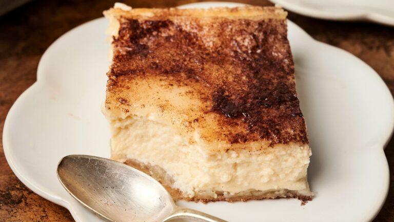 A piece of creamy cheesecake with a dusting of cocoa powder on a white plate, accompanied by a silver spoon.