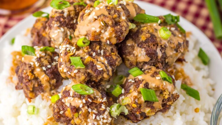 A plate of meatballs topped with sauce, sesame seeds, and chopped green onions on a bed of white rice.