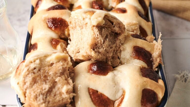 A tray of golden-brown hot cross buns with a broken piece revealing the soft interior.