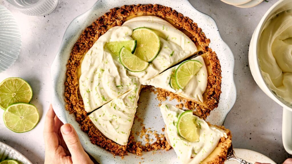 A hand reaching for a sliced key lime pie with cream topping and lime wedges, placed on a white plate. Pie crust is crumbled.