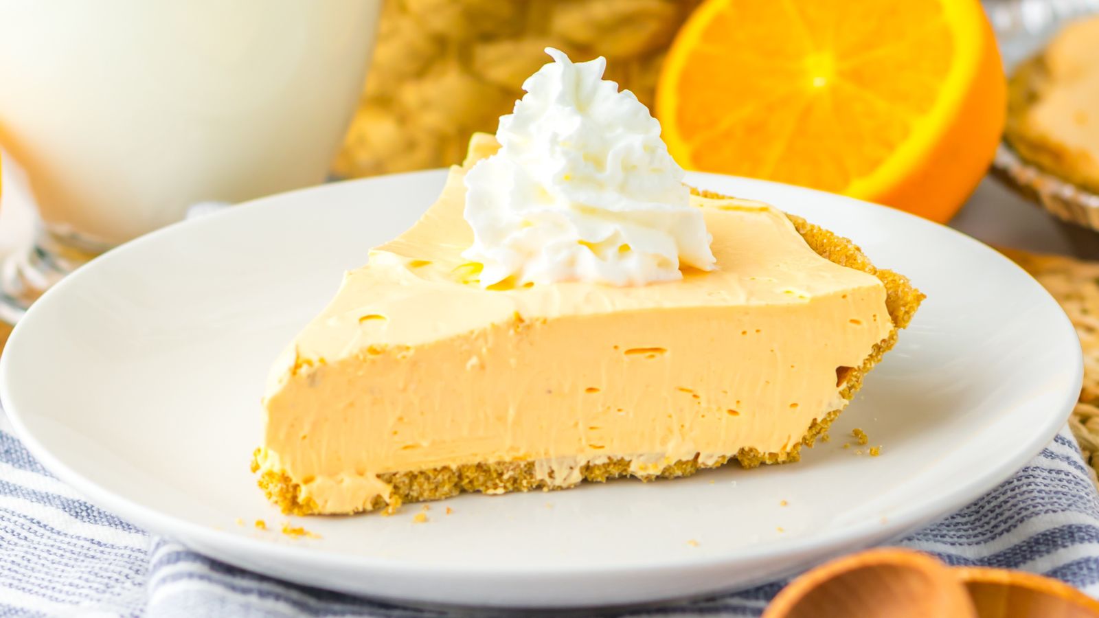A slice of cream pie on a white plate, topped with whipped cream. An orange slice and a drink are in the background.