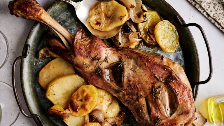 A roasted leg of lamb on a round metal tray, garnished with sliced potatoes and onions, and seasoned with rosemary. A knife is placed beside the lamb.