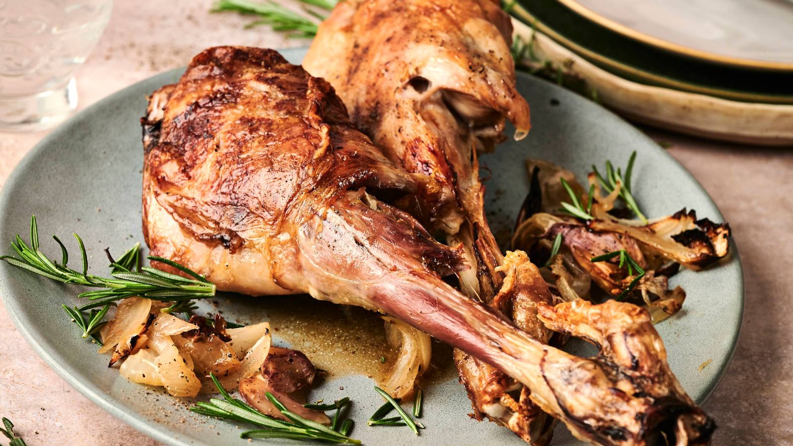 Two roasted lamb shanks on a plate with rosemary and caramelized onions, with stacked plates in the background.