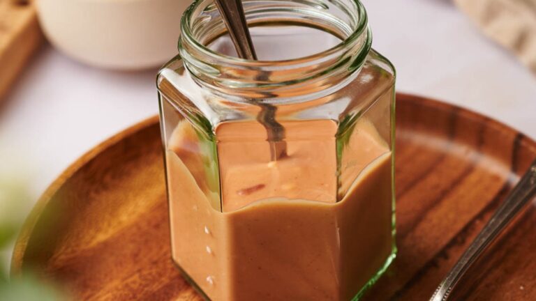 A hexagonal glass jar filled with creamy peanut butter on a wooden plate, with a metal spoon inside the jar.