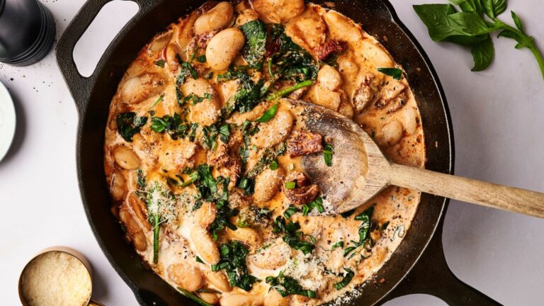 A skillet filled with creamy butter beans, wilted spinach, and sun-dried tomatoes, topped with grated cheese, with a wooden spoon resting in it.