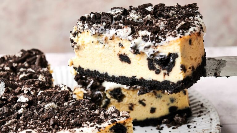 A slice of cheesecake with a cookie crumb crust and topping, featuring a creamy filling mixed with chocolate cookie pieces.