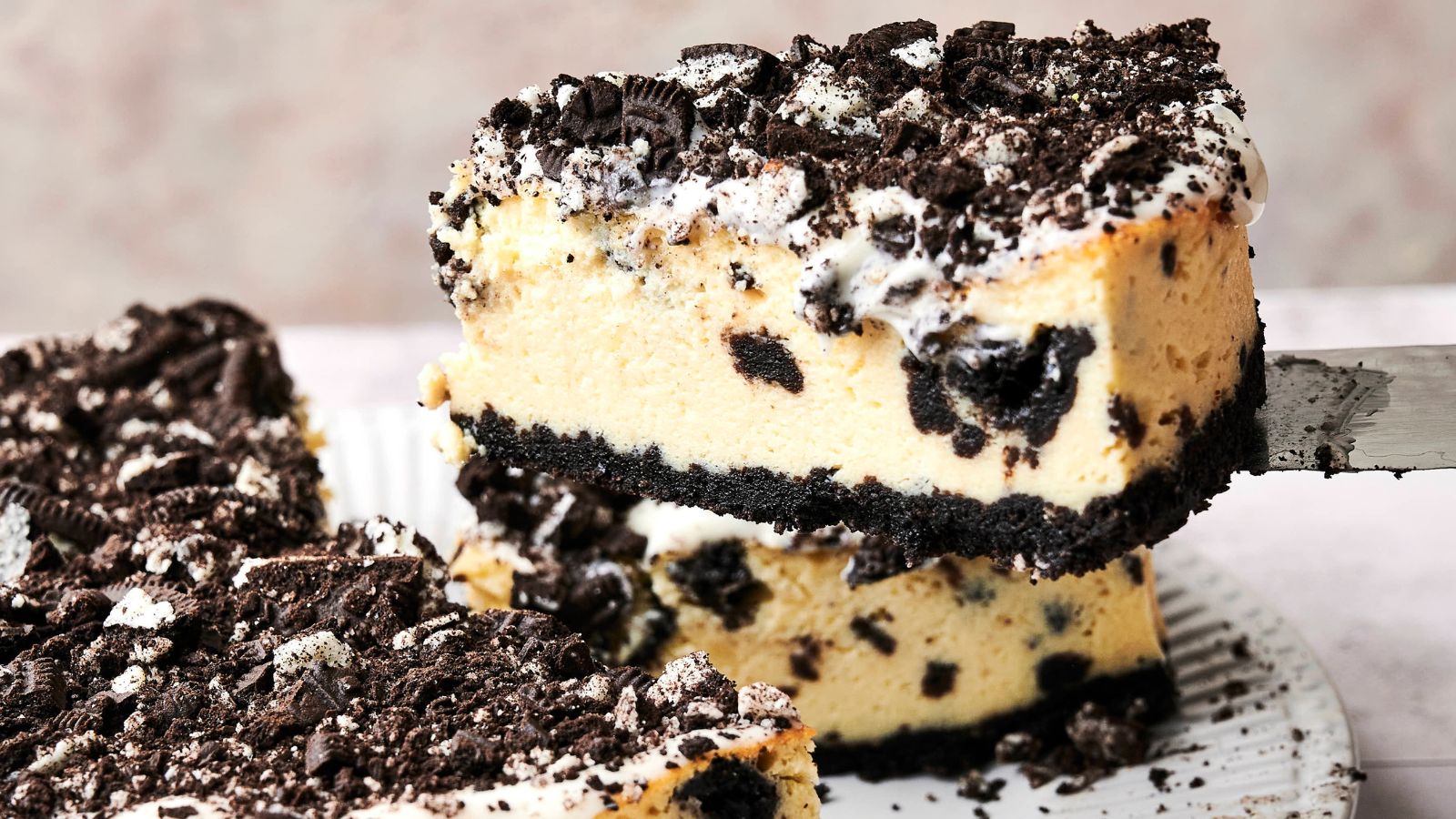 A slice of cookies and cream cheesecake is being lifted from a plate. The cake has a crumbly cookie crust, creamy filling with cookie pieces, and a topping of crushed cookies.