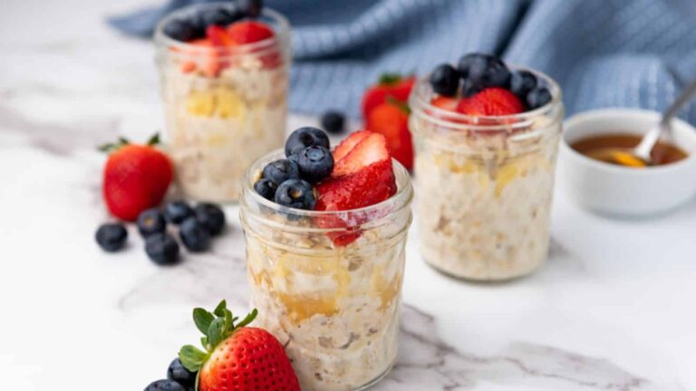 Three jars of overnight oats topped with blueberries and sliced strawberries on a marble surface, surrounded by loose berries and a small bowl of syrup.