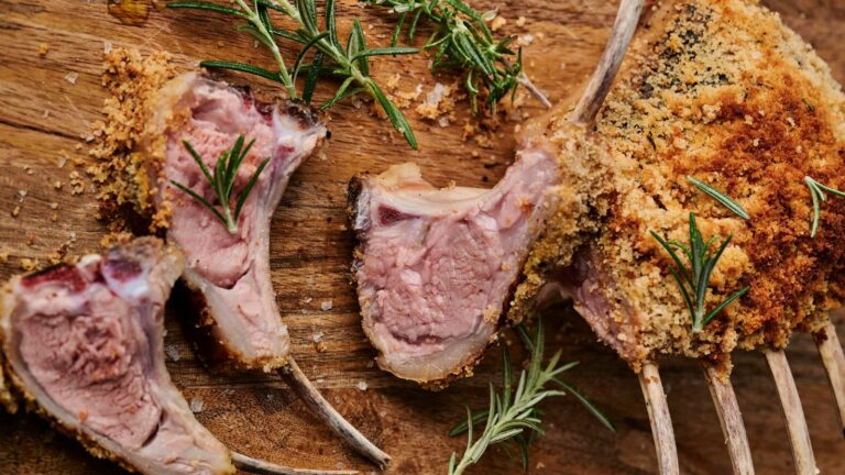 Cooked rack of lamb with a crispy breadcrumb crust, garnished with fresh rosemary, on a wooden cutting board.