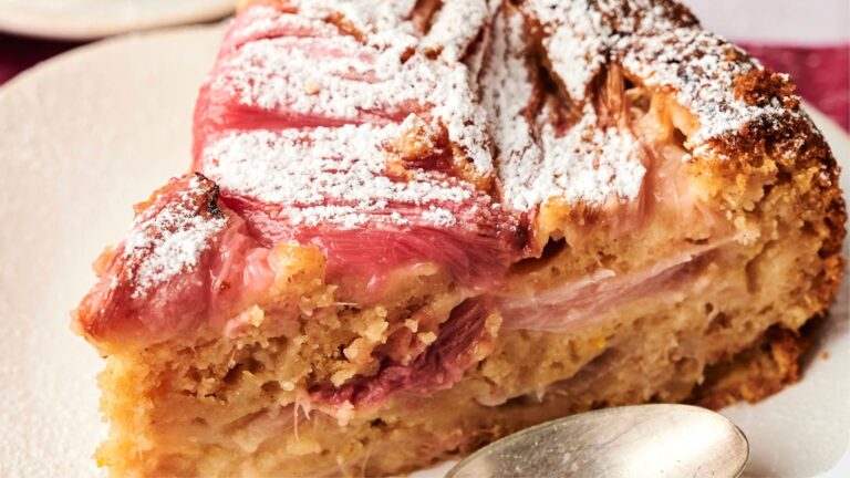 A slice of rhubarb cake topped with powdered sugar on a plate with a silver spoon beside it.