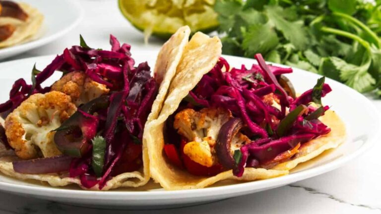 Two tacos filled with roasted cauliflower, red cabbage, and vegetables on a white plate, with cilantro and lime in the background.