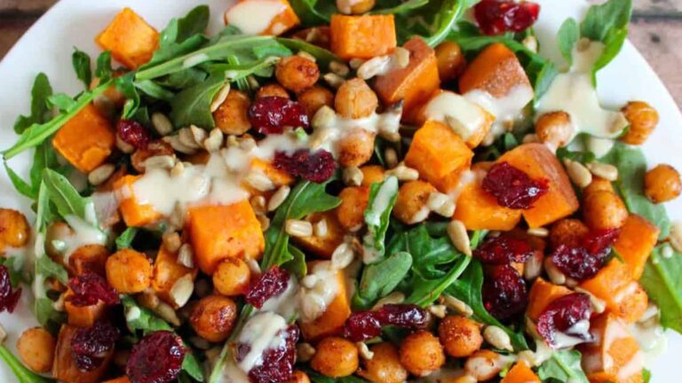 A salad with arugula, roasted chickpeas, sweet potato chunks, dried cranberries, sunflower seeds, and a drizzle of creamy dressing on a white plate.