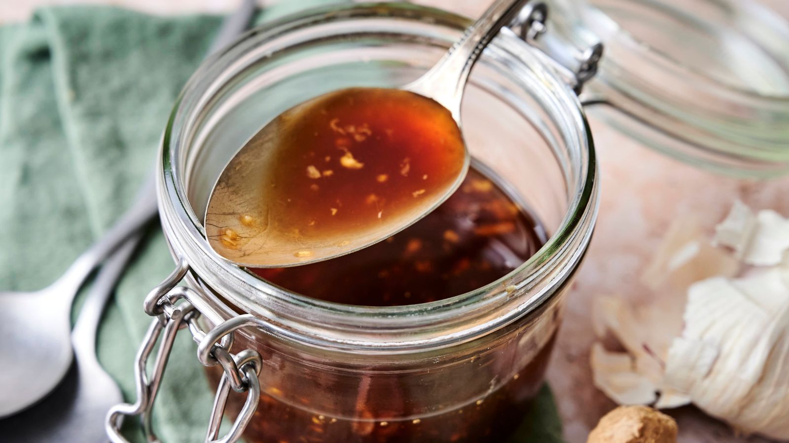 A spoon of brown liquid sauce with garlic bits is held above an open glass jar. A green cloth and garlic cloves are in the background.