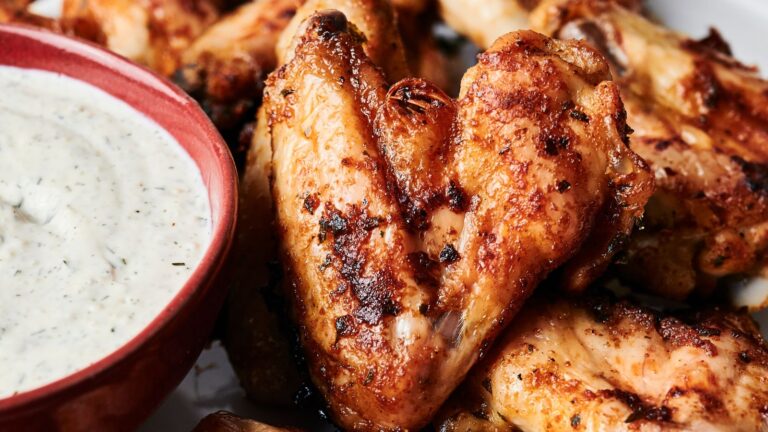 Close-up of grilled chicken wings with a crispy, seasoned exterior next to a red bowl of creamy dipping sauce.