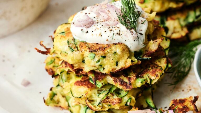 Stack of zucchini fritters topped with sour cream, chopped shallots, and dill, garnished with black pepper, on a white plate.