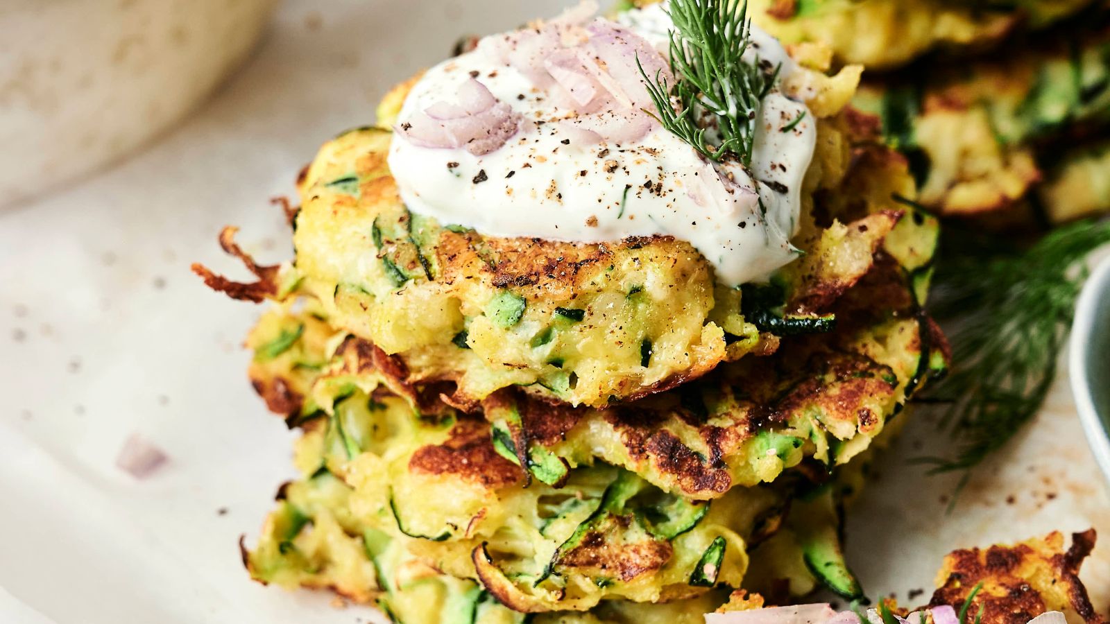 A stack of zucchini fritters topped with sour cream, thinly sliced shallots, and a sprig of dill.