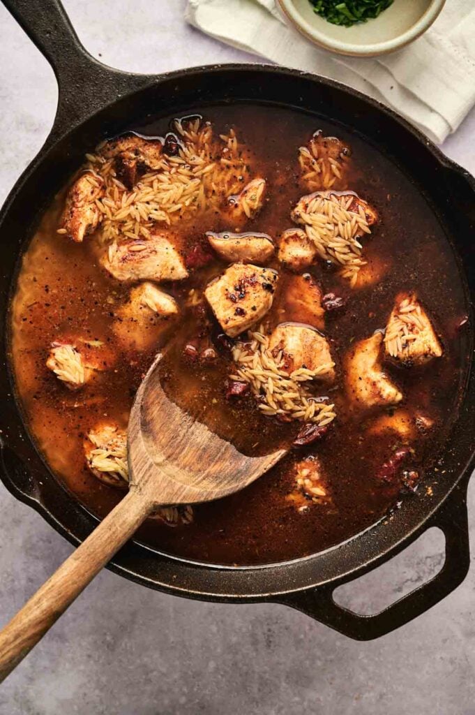 A cast iron skillet brimming with Marry Me Chicken and orzo, combined with tender rice in a rich brown sauce, all gently stirred by a wooden spoon for that perfect blend of flavors.