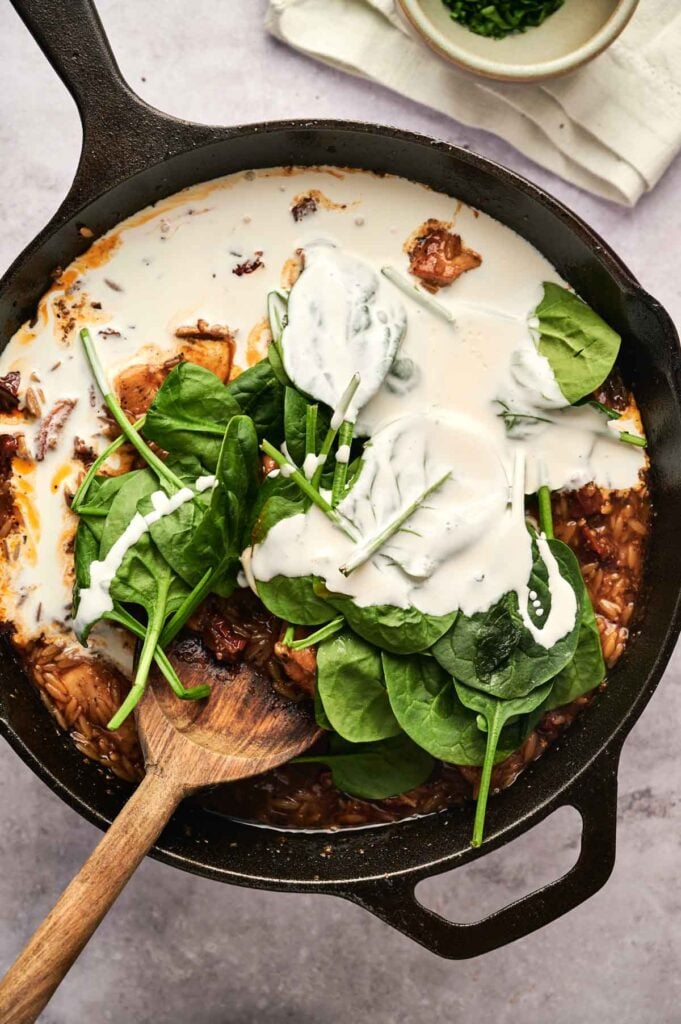 A skillet filled with creamy Marry Me Chicken Orzo, spinach, and a wooden spoon resting beside it.
