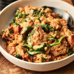 This creamy pasta dish with chicken, spinach, and tomatoes evokes the irresistible allure of Marry Me Chicken Orzo, beautifully garnished with herbs and served in a white bowl on a rustic wooden table.
