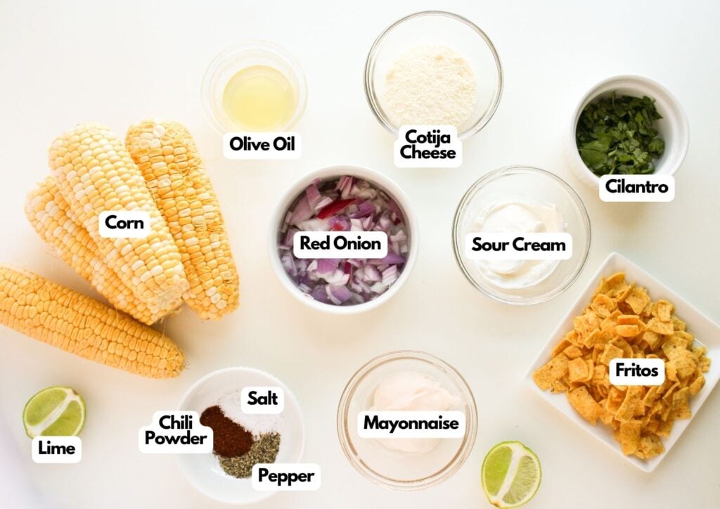 Various ingredients for a Mexican street corn salad recipe are arranged on a white surface, including corn, lime, red onion, Cotija cheese, olive oil, cilantro, sour cream, mayonnaise, Fritos, chili powder, salt, and pepper.