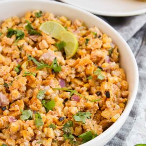 A bowl of elote salad, inspired by Mexican street corn, is garnished with lime slices and cilantro. Grilled corn sits temptingly on a plate in the background.