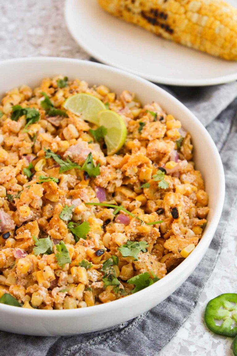 A bowl of elote salad, inspired by Mexican street corn, is garnished with lime slices and cilantro. Grilled corn sits temptingly on a plate in the background.