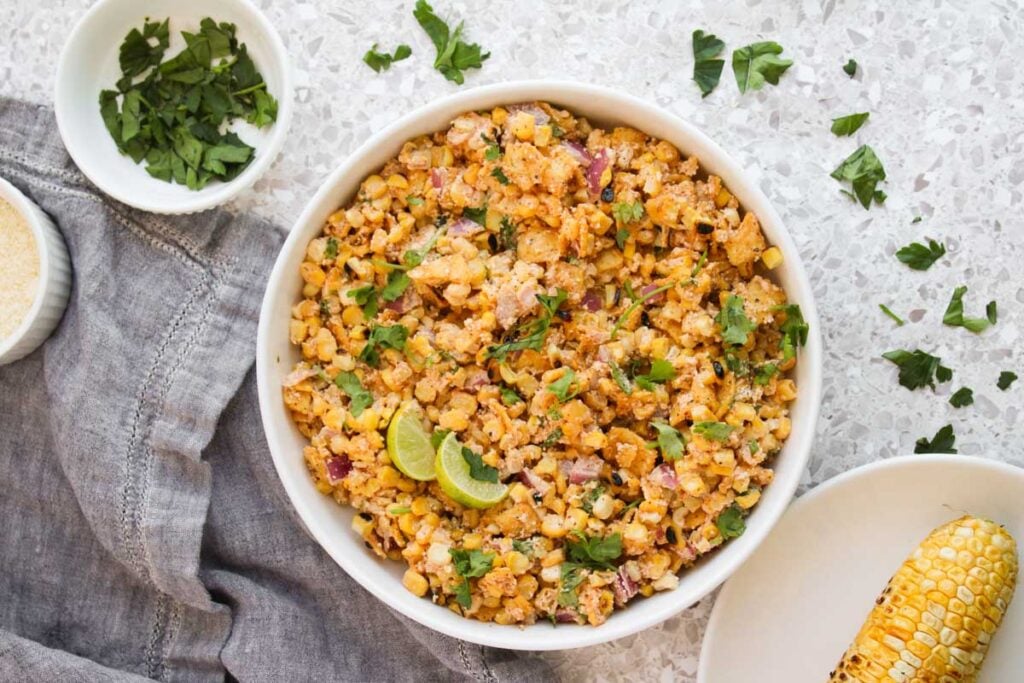 A white bowl filled with Mexican street corn salad, garnished with cilantro and lime wedges, sits on a light textured surface. Nearby are a small bowl with herbs, a corn cob, and a gray cloth.
