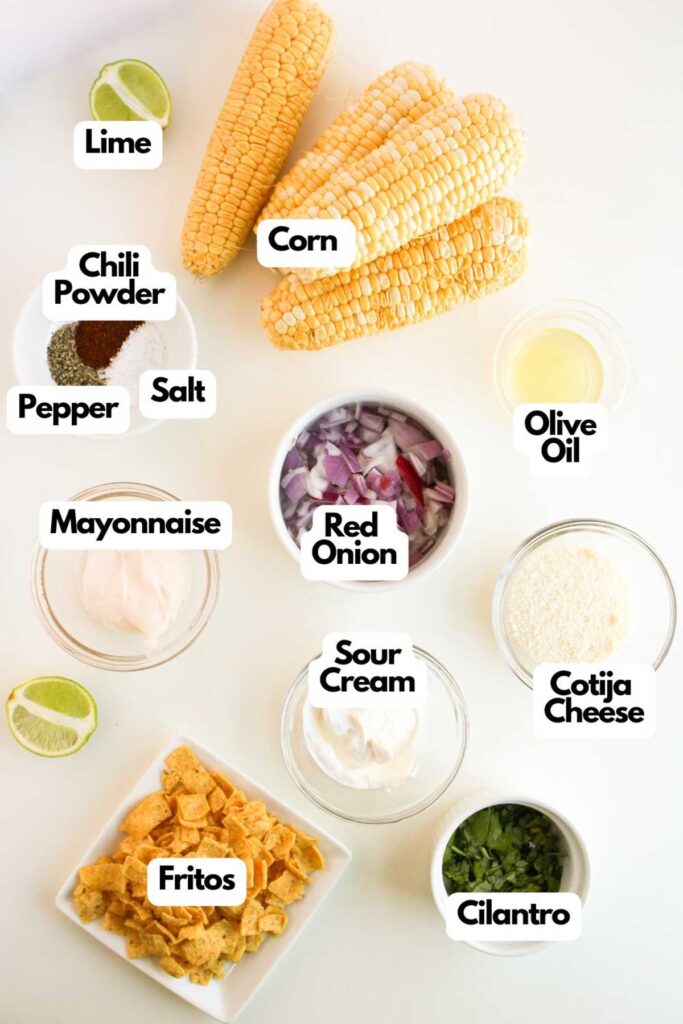 Ingredients for a vibrant Mexican street corn salad include lime, corn, chili powder, pepper, salt, olive oil, red onion, mayonnaise, sour cream, Cotija cheese, Fritos, and fresh cilantro.