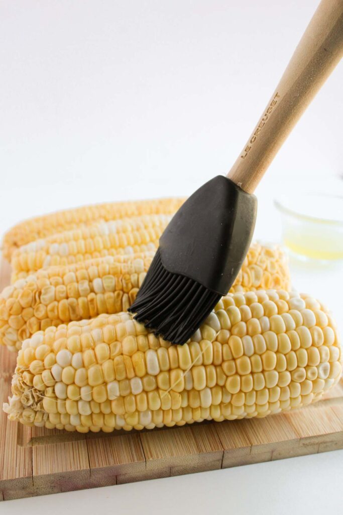 A silicone brush delicately slathers butter on ears of Mexican street corn, all atop a rustic wooden cutting board.
