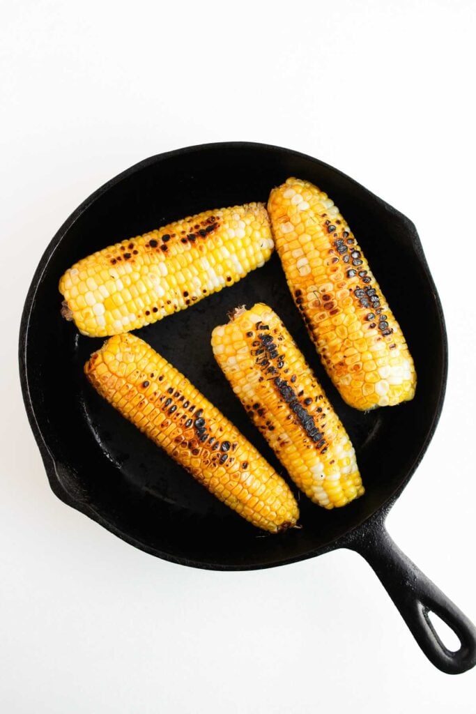 Four grilled corn cobs with char marks rest in a black cast iron skillet, capturing the essence of Mexican street corn on a white background.