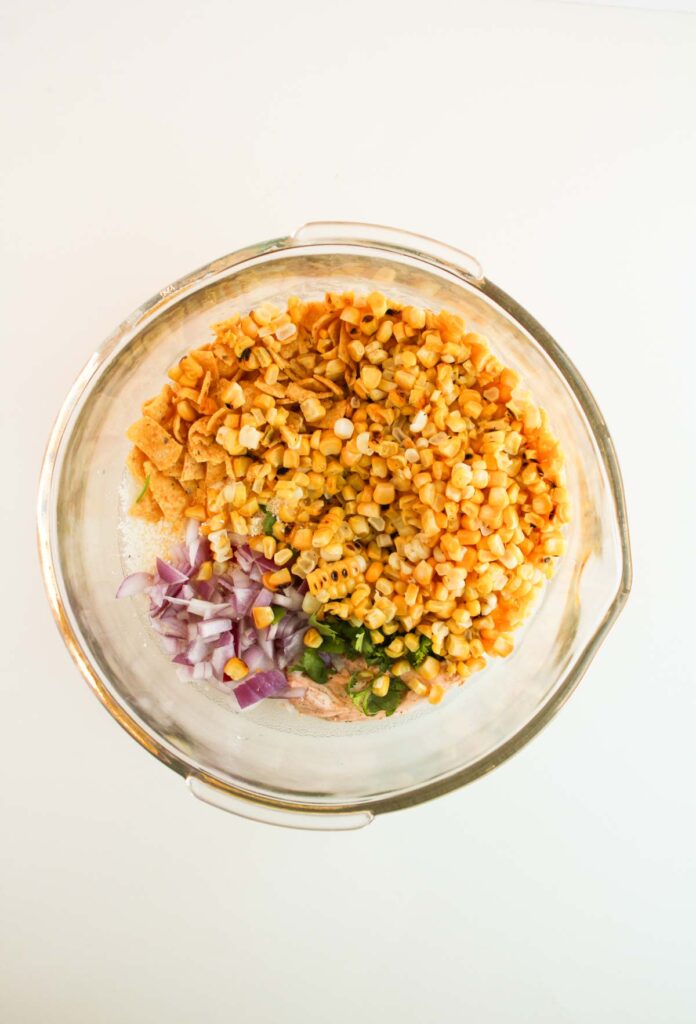 A glass bowl containing chopped red onion, jalape&ntilde;os, corn, and broken tortilla chips sits on a white surface, reminiscent of a vibrant Mexican street corn medley.