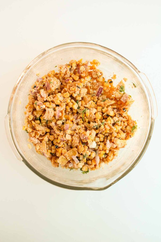 A clear glass bowl filled with a vibrant mixture reminiscent of Mexican street corn, featuring crushed tortilla chips, diced vegetables, and herbs on a pristine white surface.